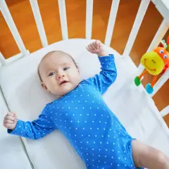 Urocze, 10 tygodniowe dziecko w niebieskim pajacyku leży w łóżeczku obok kolorowej zabawki.
