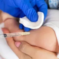 Ręka w niebieskiej rękawiczce wykonuje szczepienia noworodka. Wacik i strzykawka są gotowe do podania leku.