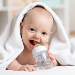 Uśmiechnięte niemowlę pod białym ręcznikiem pije wodę dla niemowląt z butelki.