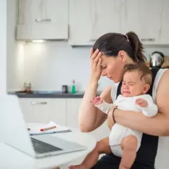 Zmęczona mama z płaczącym, **high need baby** na rękach, próbuje pracować przy laptopie.
