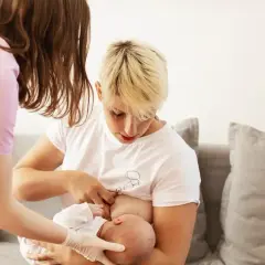 Doradca laktacyjny pomaga mamie w karmieniu piersią. Na zdjęciu widać kobietę w białej koszulce karmiącą dziecko, a obok niej drugą kobietę w różowej bluzce.