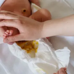 Rączka rodzica delikatnie dotyka stópki niemowlaka, podczas gdy pieluszka pokazuje zawartość – kupa niemowlaka.