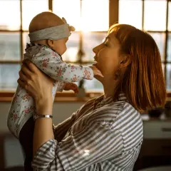 Mama bawi się z dzieckiem, które wykonuje odginanie głowy do tyłu. Ciepłe światło zachodzącego słońca oświetla ich twarze.