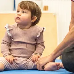 Małe dziecko siedzi na macie, a dorosły trzyma je za stopę. Czy 5 miesięczne dziecko można sadzać na kolanach?