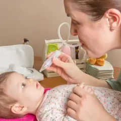 Mama pomaga noworodkowi z katarem, używając aspiratora. Dziecko leży na różowym ręczniku, patrząc na mamę.