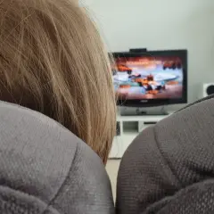 Niemowlę z blond włosami siedzi na kanapie, a w tle widać włączony telewizor z grą.
