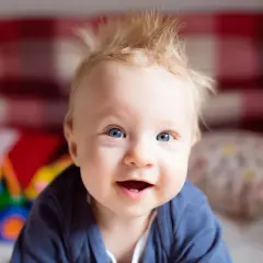 Uśmiechnięte niemowlę z blond czuprynką, któremu jakby nie rosną włosy. Ma niebieskie oczy i wesołe spojrzenie.