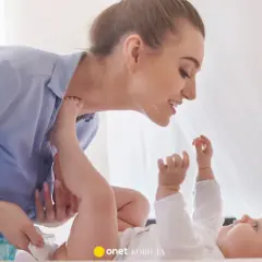 Uśmiechnięta mama pochyla się nad dzieckiem, które leży na plecach. Wykonuje szerokie pieluchowanie, delikatnie unosząc nóżki malucha.