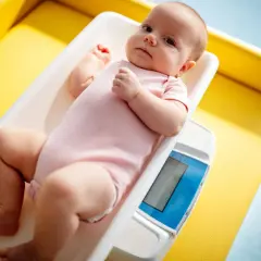 Noworodek w różowym body leży na wadze. Rodzice często zastanawiają się, ile powinien przybierać noworodek, by mieć pewność, że rozwija się prawidłowo.