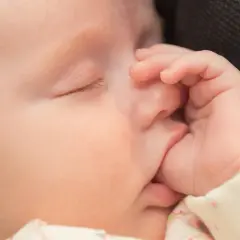 Uroczy niemowlak śpi, a jego mała rączka delikatnie obejmuje usta. To naturalne ssanie kciuka u niemowlaka, które przynosi mu ukojenie.