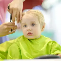 Mały chłopiec z blond włosami przeżywa swoje pierwsze strzyżenie dziecka. Fryzjerka delikatnie podcina mu włosy.