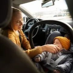 Uśmiechnięta mama przygotowuje dziecko do podróży. Podpowiadamy, jak ubrać dziecko do fotelika zimą, by było mu ciepło i bezpiecznie.