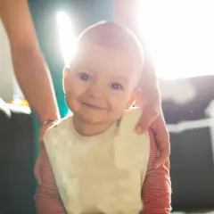 Uśmiechnięte niemowlę z pianą z ust, w śliniaczku, otoczone rękami opiekunów.