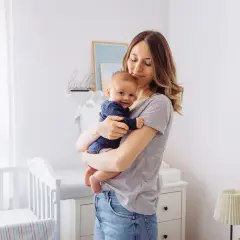 Uśmiechnięty, 2 miesięczne dziecko w ramionach mamy. W tle pokój dziecięcy z łóżeczkiem.
