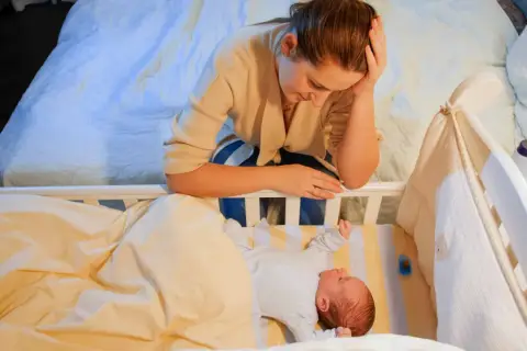 Zmęczona mama pochyla się nad łóżeczkiem, zastanawiając się, kiedy nastąpi regres snu. Niemowlę leży spokojnie pod kołderką.