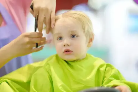 Mały chłopiec z blond włosami przeżywa swoje pierwsze strzyżenie dziecka. Fryzjerka delikatnie podcina mu włosy.