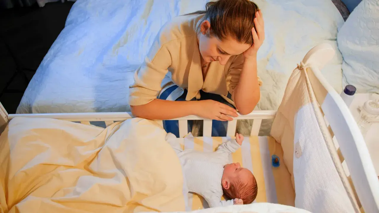 Zmęczona mama pochyla się nad łóżeczkiem, zastanawiając się, kiedy nastąpi regres snu. Niemowlę leży spokojnie pod kołderką.