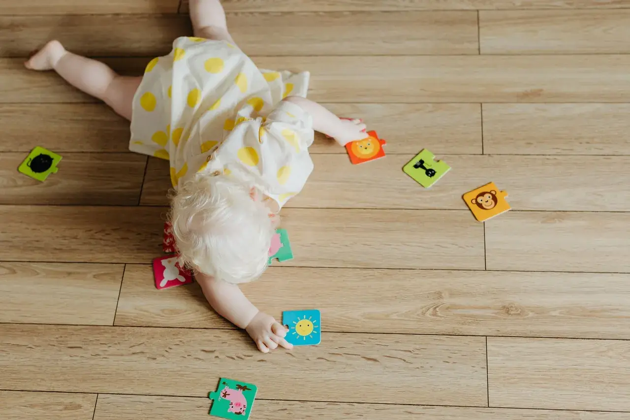 Dziecko z jasnymi włosami, w żółtej sukience w kropki, bawi się na podłodze. Rozrzucone są puzzle ze zwierzątkami i słońcem. To moment, kiedy dziecko zaczyna się obracać.