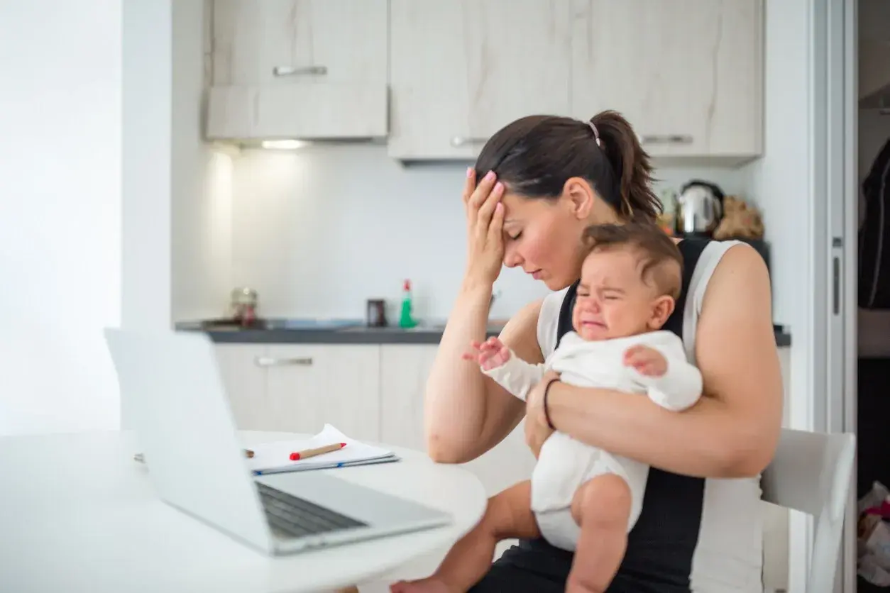 Zmęczona mama z płaczącym, **high need baby** na rękach, próbuje pracować przy laptopie.