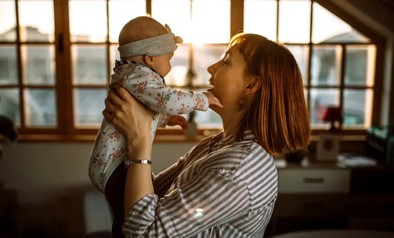 Mama bawi się z dzieckiem, które wykonuje odginanie głowy do tyłu. Ciepłe światło zachodzącego słońca oświetla ich twarze.