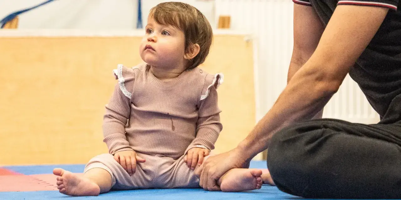 Małe dziecko siedzi na macie, a dorosły trzyma je za stopę. Czy 5 miesięczne dziecko można sadzać na kolanach?