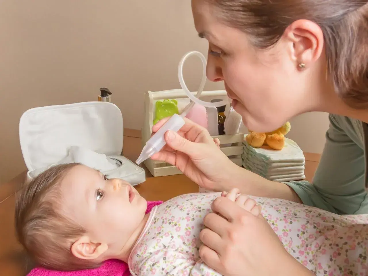 Mama pomaga noworodkowi z katarem, używając aspiratora. Dziecko leży na różowym ręczniku, patrząc na mamę.
