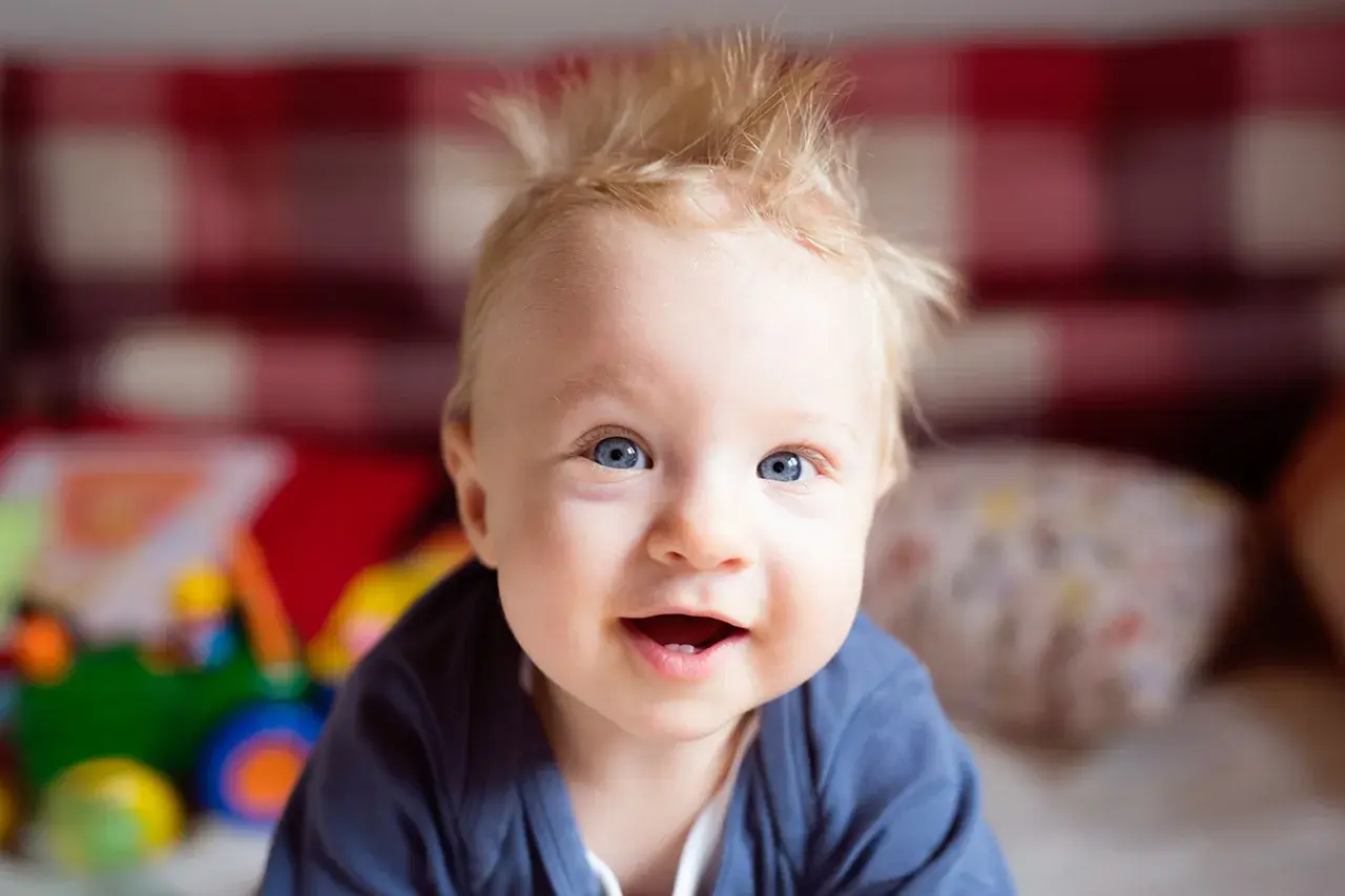 Uśmiechnięte niemowlę z blond czuprynką, któremu jakby nie rosną włosy. Ma niebieskie oczy i wesołe spojrzenie.