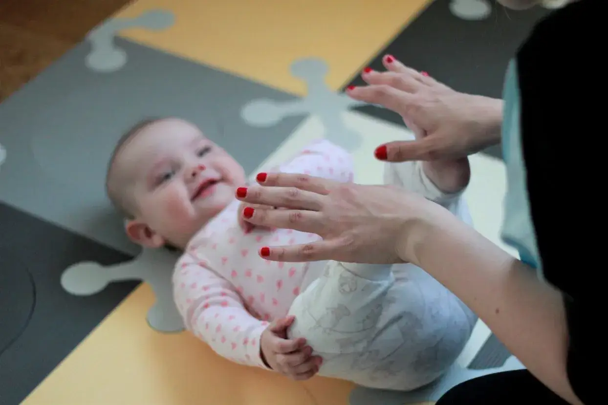 Uśmiechnięte dziecko na macie, ręce mamy z czerwonymi paznokciami dotykają jego nóżek. To czas skoku rozwojowego 6 miesiąca.