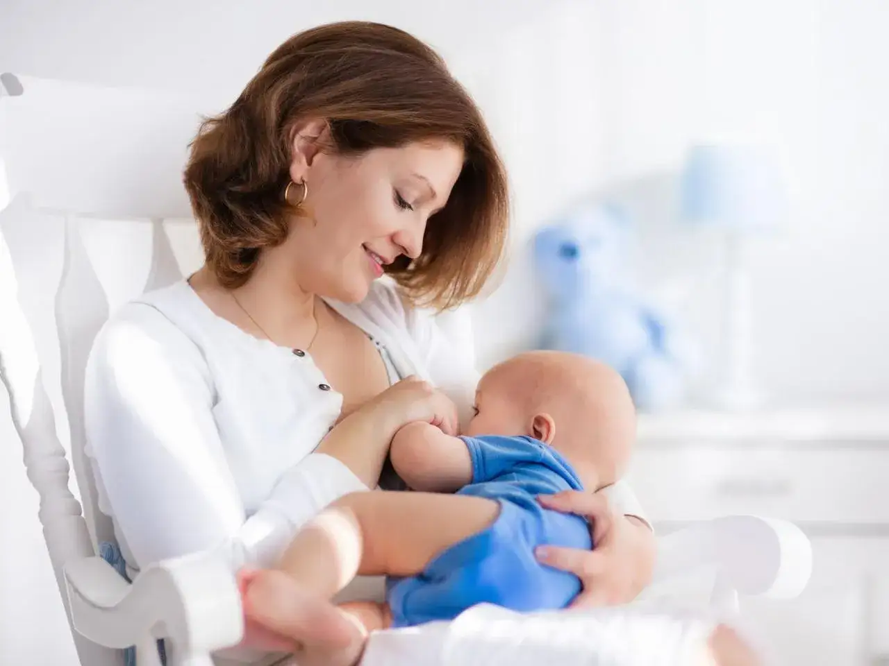 Uśmiechnięta mama karmi piersią dziecko w białym fotelu. Czy można jeść miód karmiąc piersią? To pytanie nurtuje wiele mam.