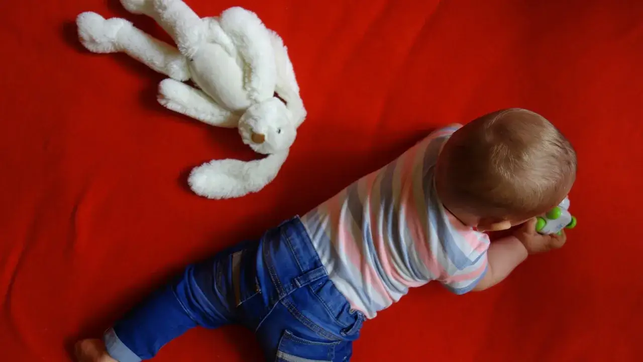 Małe dziecko w dżinsach i pasiastym body bawi się zabawką, kiedy dziecko raczkuje. Obok leży biały pluszowy króliczek.