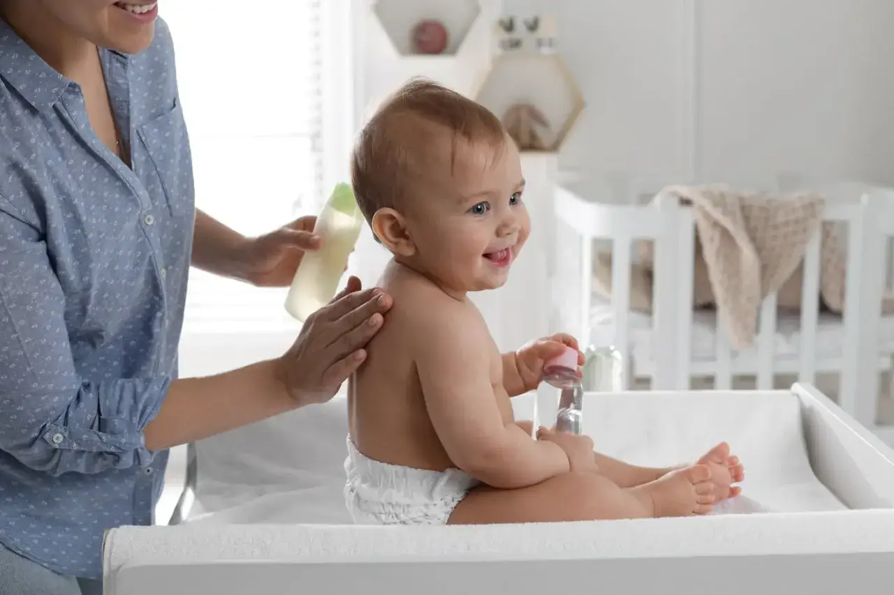 Mama delikatnie wmasowuje w plecy dziecka olej ze słodkich migdałów. Uśmiechnięty noworodek trzyma w rączce buteleczkę z płynem.