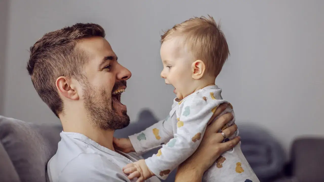 Ojciec śmieje się do swojego high need baby, który wyciąga rączki. Radość i bliskość w tej chwili.