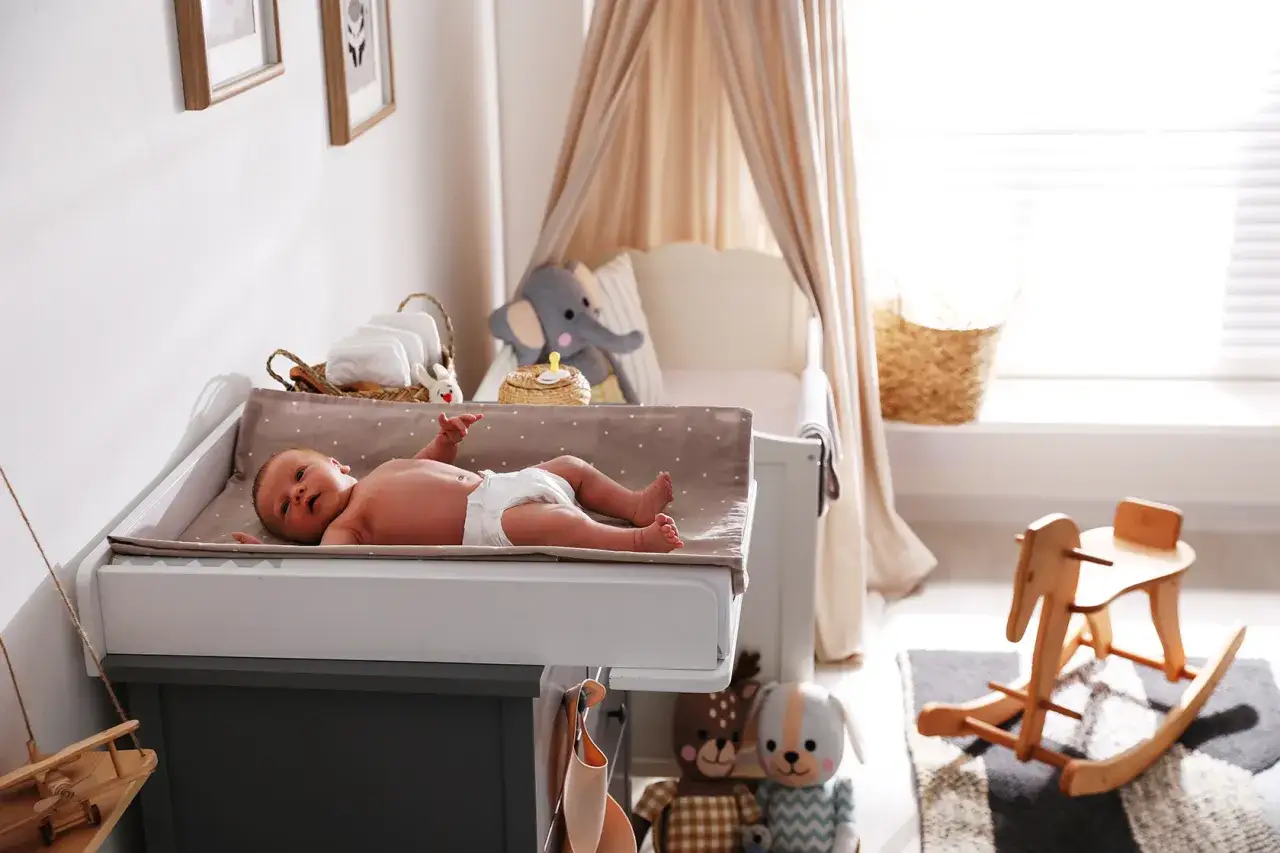 Niemowlę na przewijaku, obok koszyk z pieluchami i pluszowy słoń. W tle łóżeczko i drewniany konik na biegunach. Wszystko gotowe na przyjście maluszka, nawet czopki glicerynowe dla niemowląt.