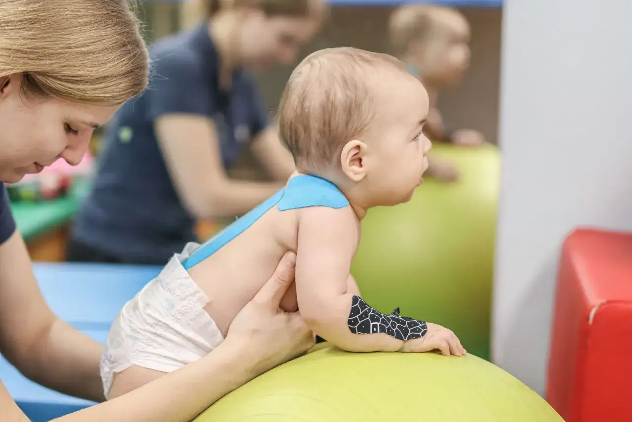 Fizjoterapeutka wspiera dziecko z wzmożonym napięciem mięśniowym na piłce rehabilitacyjnej.