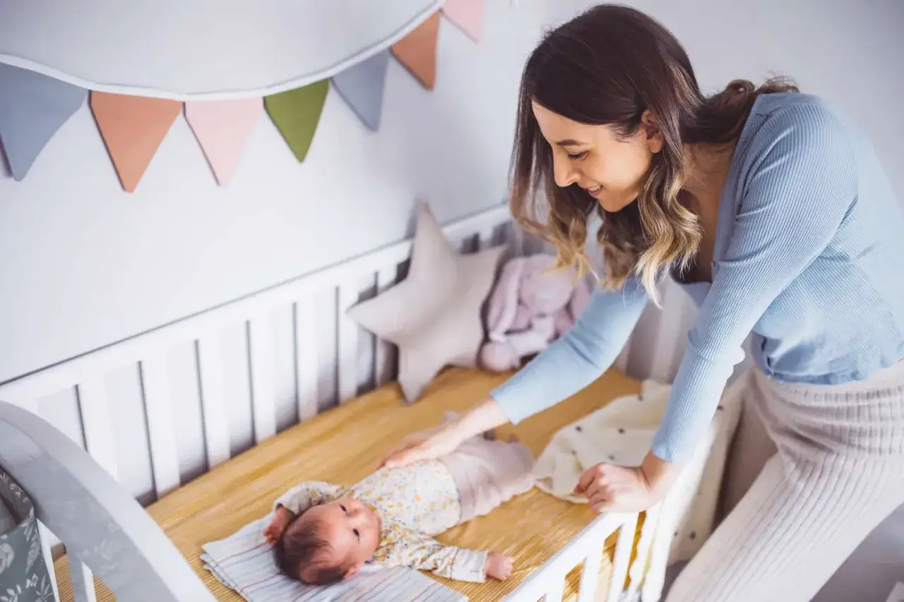 Mama czule głaszcze śpiącego noworodka w łóżeczku. Słychać ciche stękanie noworodka podczas snu, co jest naturalnym odgłosem.