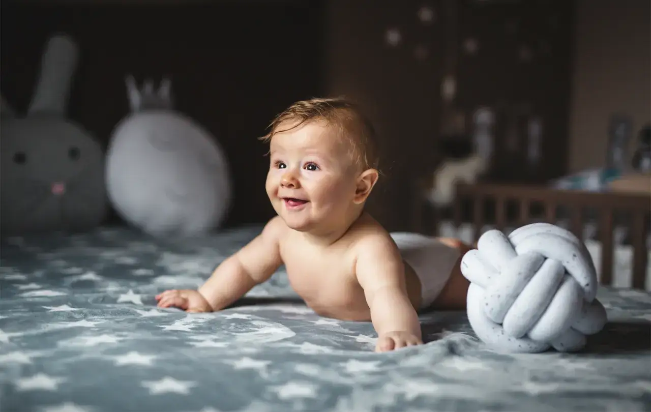 Uśmiechnięte dziecko, 22 tydzień życia, leży na kocu w gwiazdki. Obok pluszowa kula i zabawki.